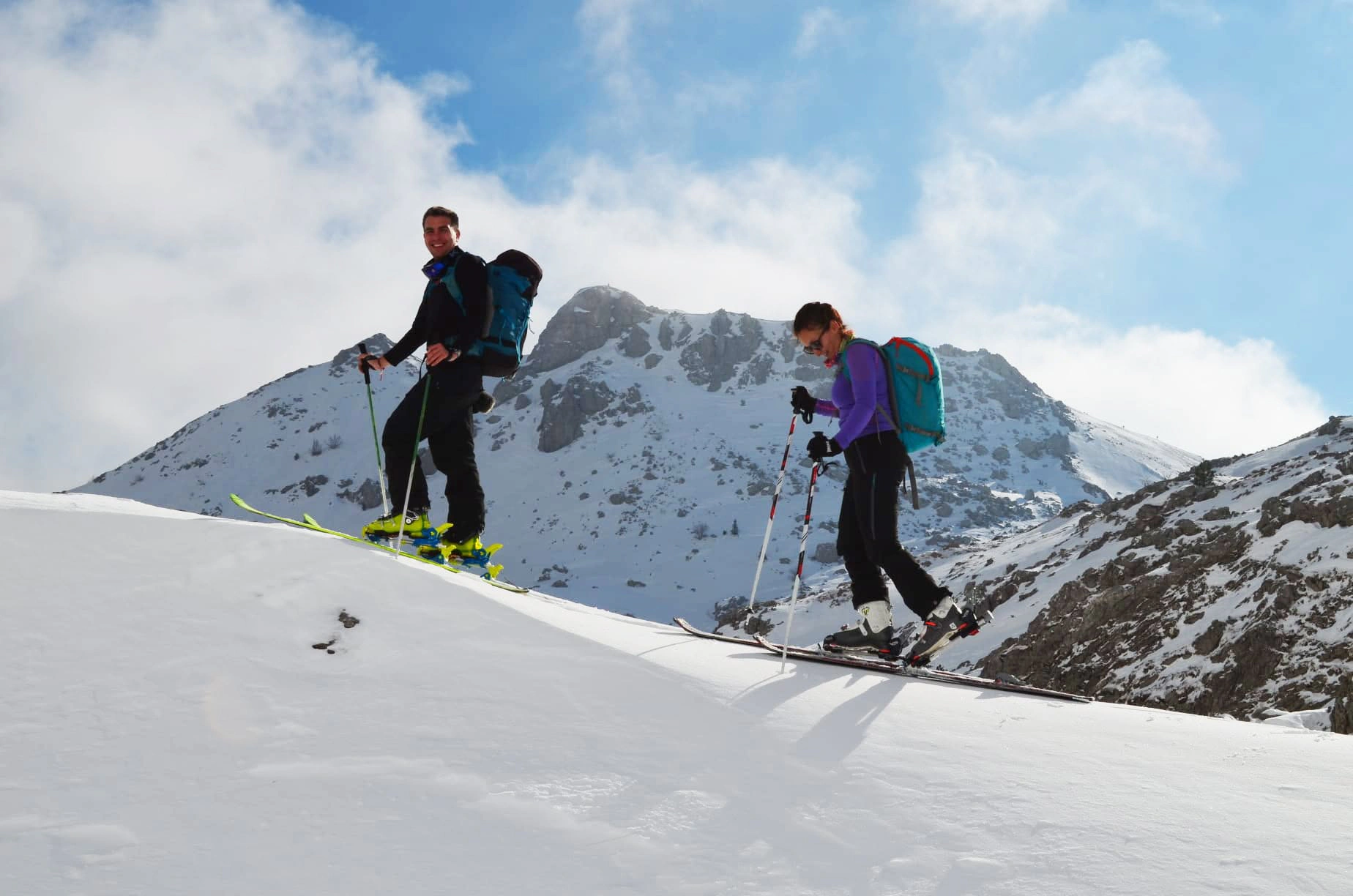 Dejan and a friend ski touring up a snowy ridge below a rocky peak